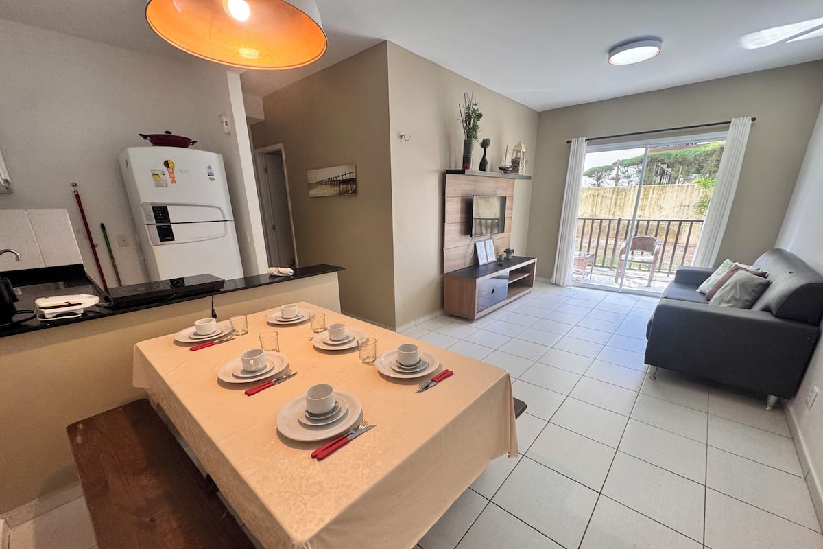 A modern kitchen with white appliances, a dining area with a table set for four, a sofa, and a balcony with a view.