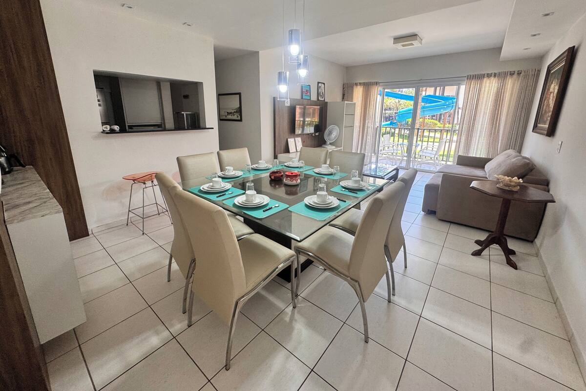 A modern dining area with a glass table set for six, surrounded by beige chairs. A kitchen area with a TV and cabinets is visible to the left.