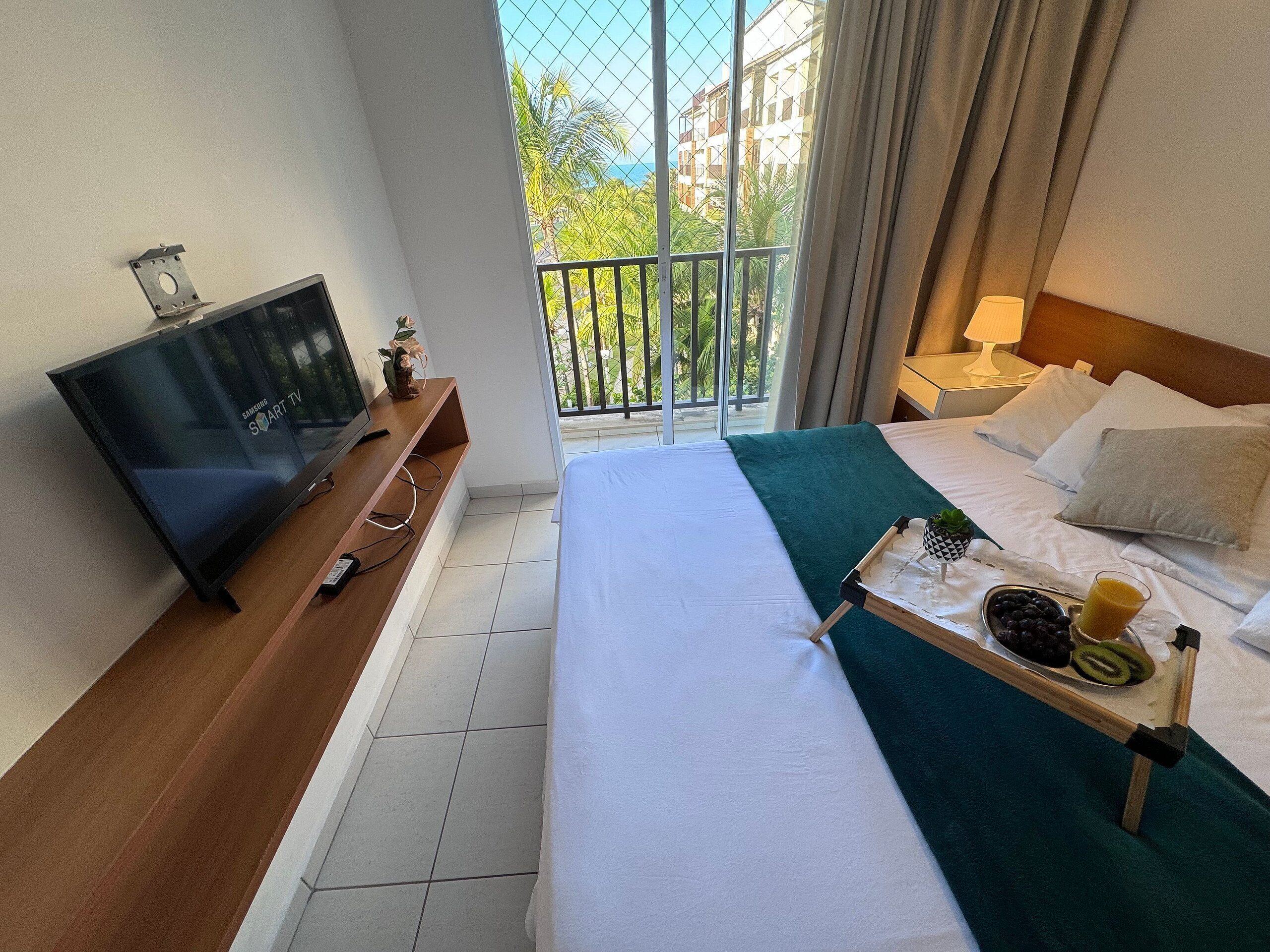 A hotel room with a bed, a TV on a wooden stand, a balcony with a view of buildings and greenery, and a tray with fruits and a glass of juice on the bed.