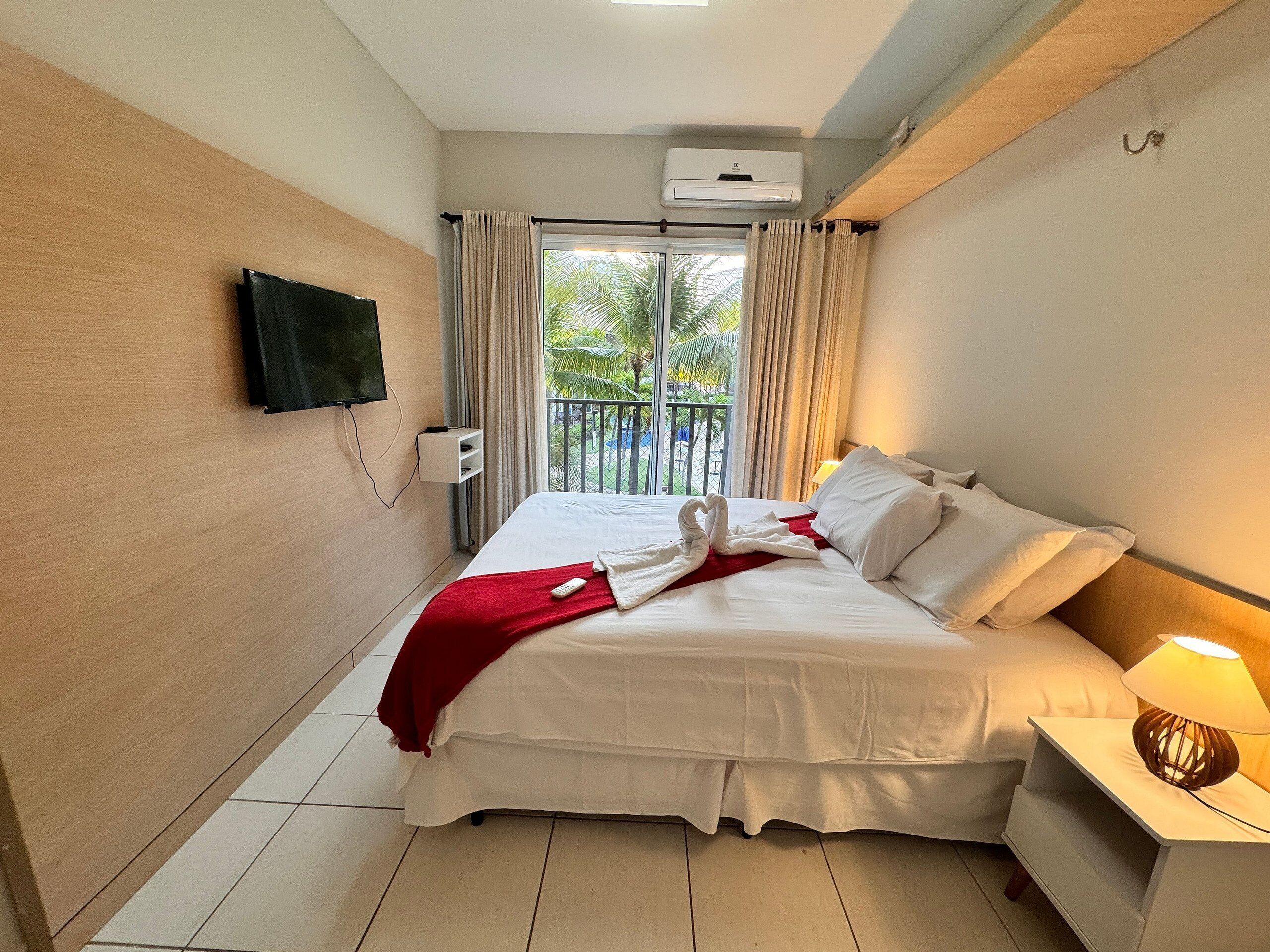 A hotel room with a bed, a TV mounted on the wall, a balcony with curtains, and an air conditioning unit.