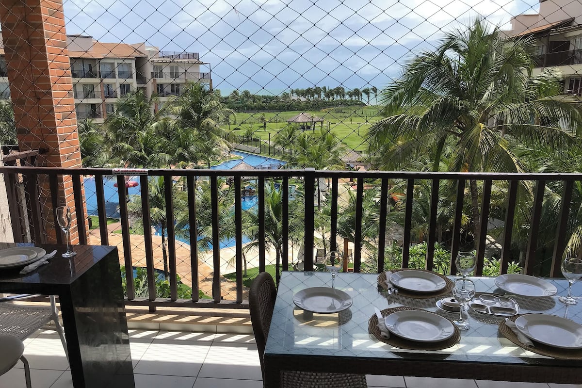 A balcony with a table set for dining, overlooking a swimming pool and a green landscape with palm trees.
