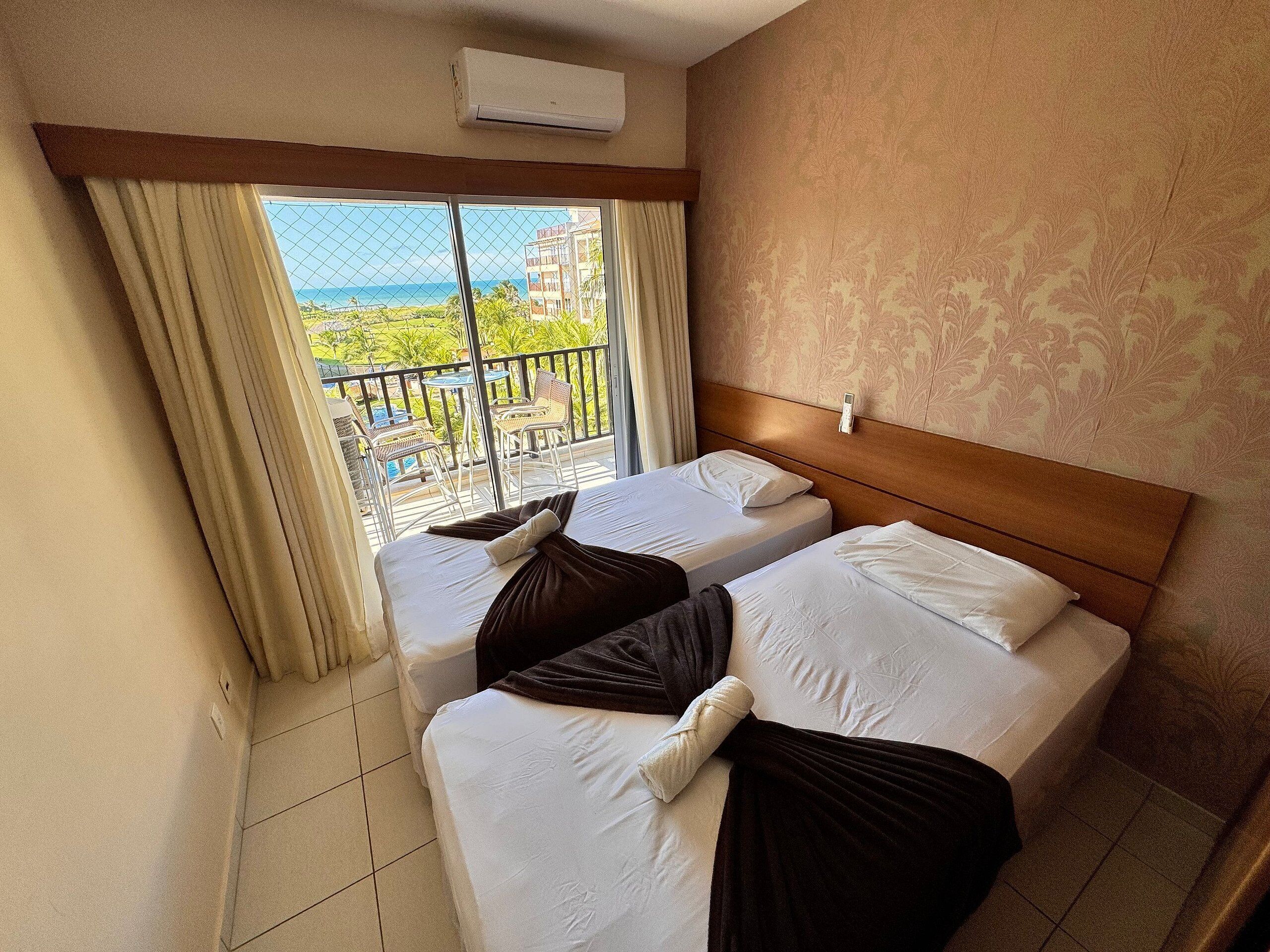 Habitació d'hotel amb dos llits, un balcó que ofereix vistes al mar i als edificis, i un aparell d'aire condicionat a la paret.