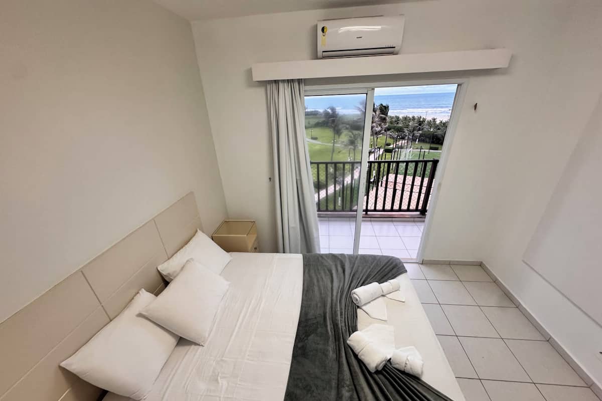 A hotel room with a bed, a nightstand, an air conditioning unit, a balcony with a view of the ocean and a golf course.