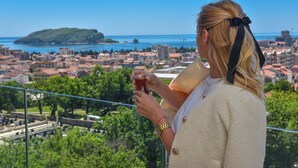 Una persona sosteniendo un vaso, con vistas a una ciudad costera con edificios y zonas verdes.