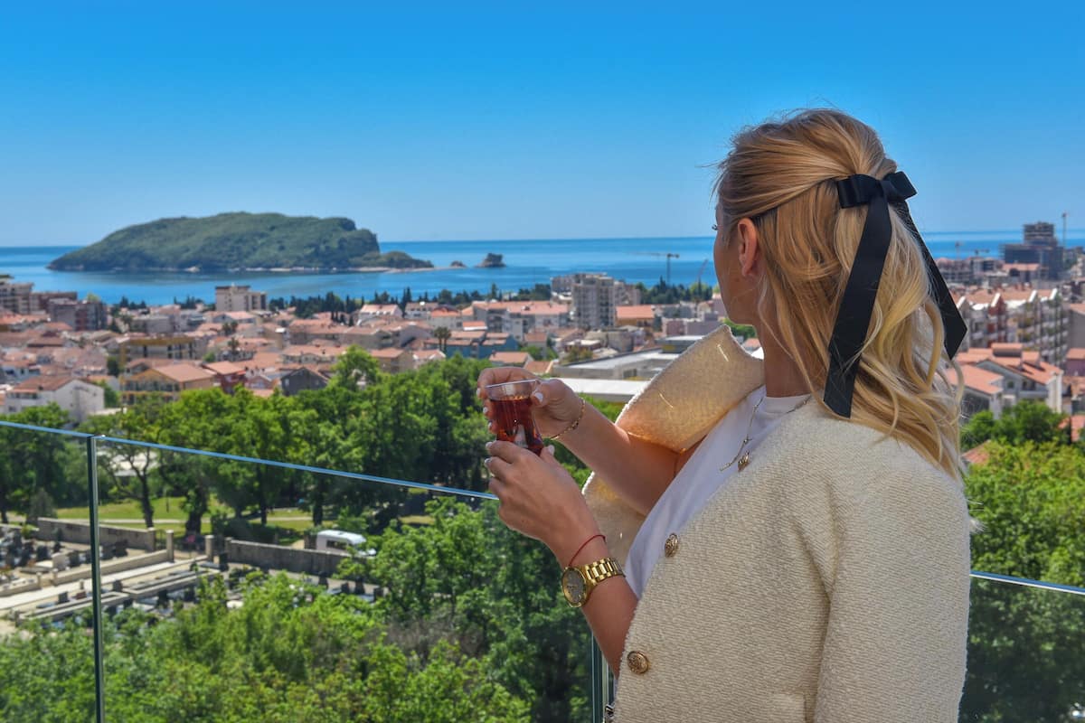 A person holding a glass of drink, overlooking a coastal cityscape with buildings and greenery.