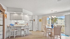A modern kitchen with white cabinets, a dining area with white chairs, and a view of a residential area through a large window. - Shorehaven II Beach Resort (North Myrtle Beach)