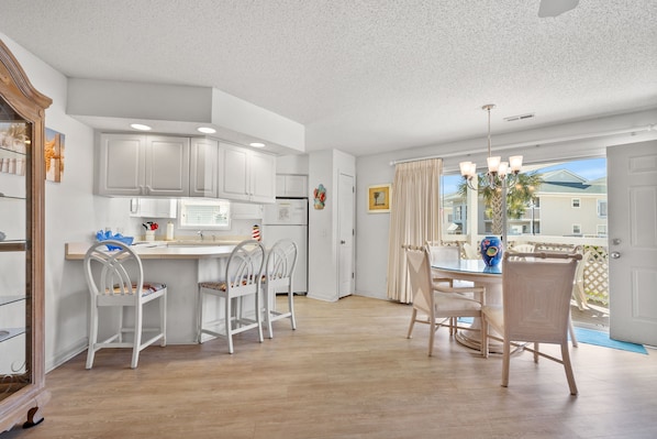 A modern kitchen with white cabinets, a dining area with white chairs, and a view of a residential area through a large window. - Shorehaven II Beach Resort (North Myrtle Beach)