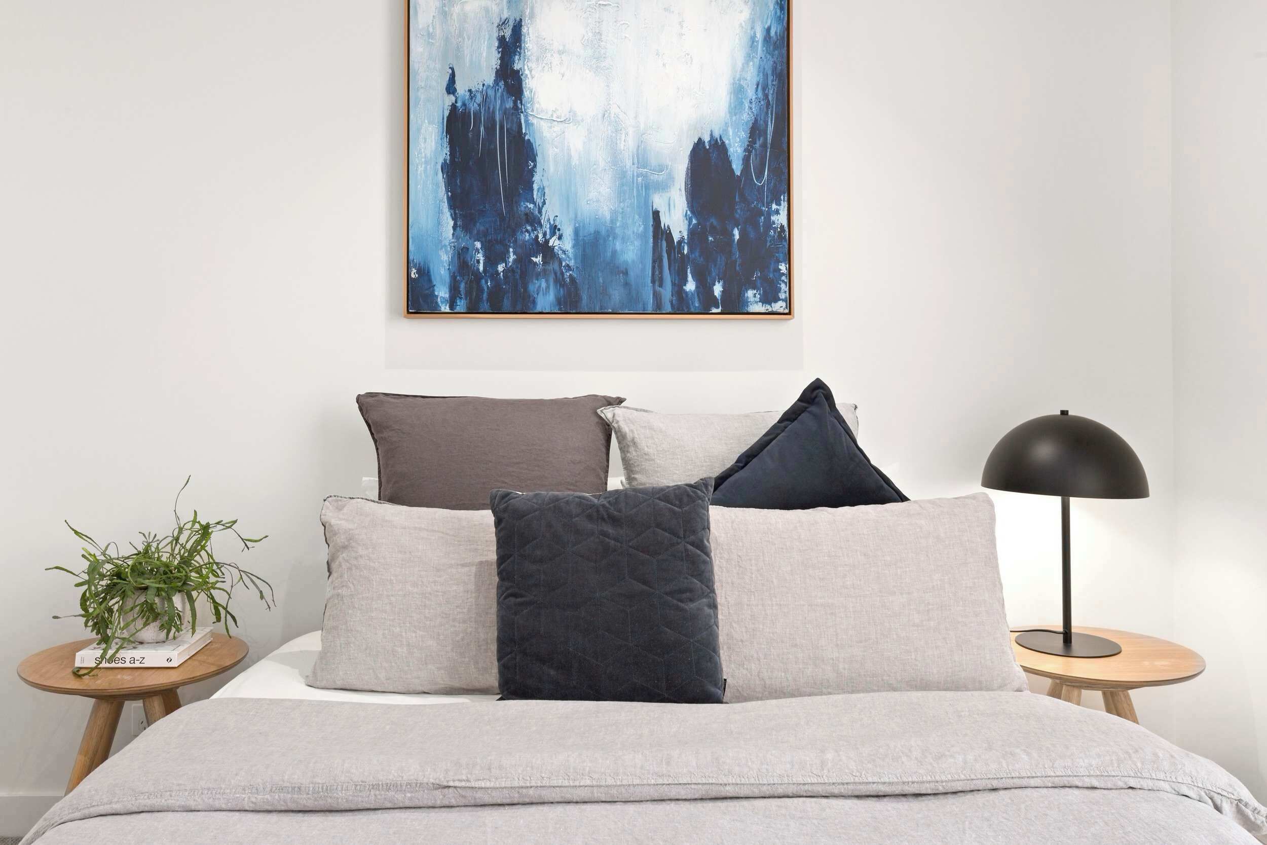 Une chambre à coucher avec un lit, des oreillers, une table de chevet avec une lampe et un tableau abstrait accroché au mur.