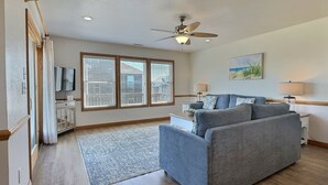 Ein Wohnzimmer mit grauem Sofa, Deckenventilator, Fernseher und einem Fenster mit Jalousien.