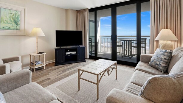 A hotel room with a balcony, a flat-screen TV, a sofa, a coffee table, and a lamp.