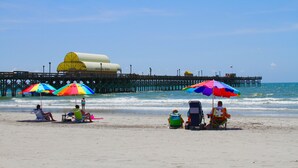 A beach with people relaxing under colorful umbrellas, a pier in the background, and the ocean. - Tilghman Lakes By the Sea (North Myrtle Beach)