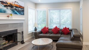A modern living room with a fireplace, a sectional sofa with red cushions, a round coffee table, and large windows with blinds. - Painted Cliff 18  Ski-in, Ski-out Luxury with serene mountain view, sleeps 7 (Whistler)