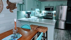A modern kitchen with teal cabinetry, stainless steel appliances, and a wooden dining table with a decorative bird centerpiece. - Sea Cloisters I Ocean Front Beach Retreat (North Myrtle Beach)