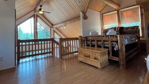 A spacious room with a high ceiling, wooden beams, a bed with patterned bedding, a chest of drawers, and large windows offering a view of trees. - 1 BR+ Loft, Log Cabin With Lakeview, Sleeps 4, 5 mins to Glacier (West Glacier)