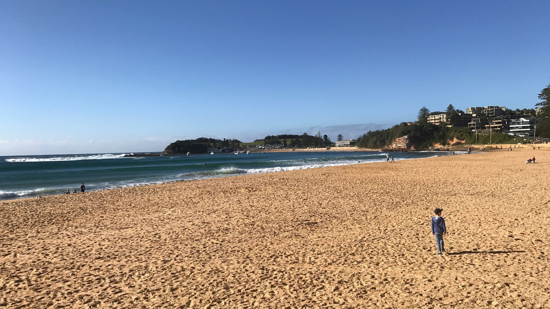 A wide sandy beach with a clear blue sky, a few people, and buildings in the background.
