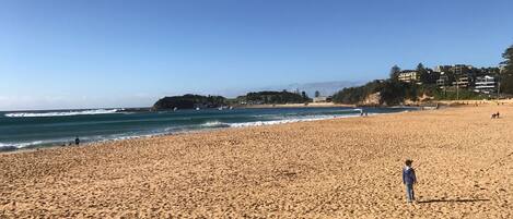A wide sandy beach with a clear blue sky, a few people, and buildings in the background.