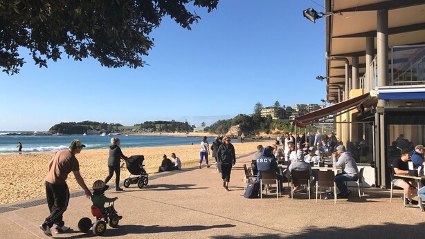 Um café à beira-mar com mesas ao ar livre, vista para a praia e céu claro.