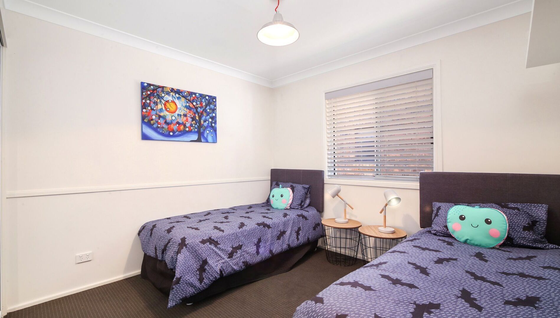 A bedroom with two beds, a window with blinds, a table with two lamps, and a colorful painting on the wall.