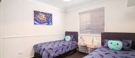 A bedroom with two beds, a window with blinds, a table with two lamps, and a colorful painting on the wall.