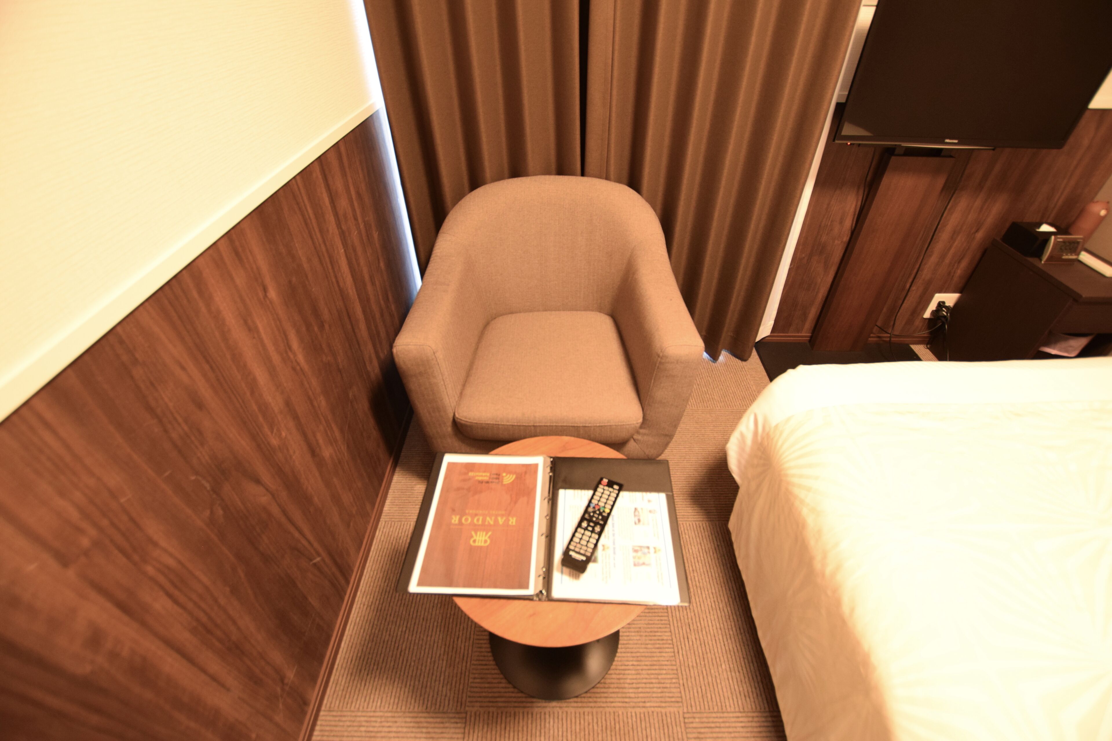 A hotel room with a bed, a chair, a small round table, a remote control, and a book.
