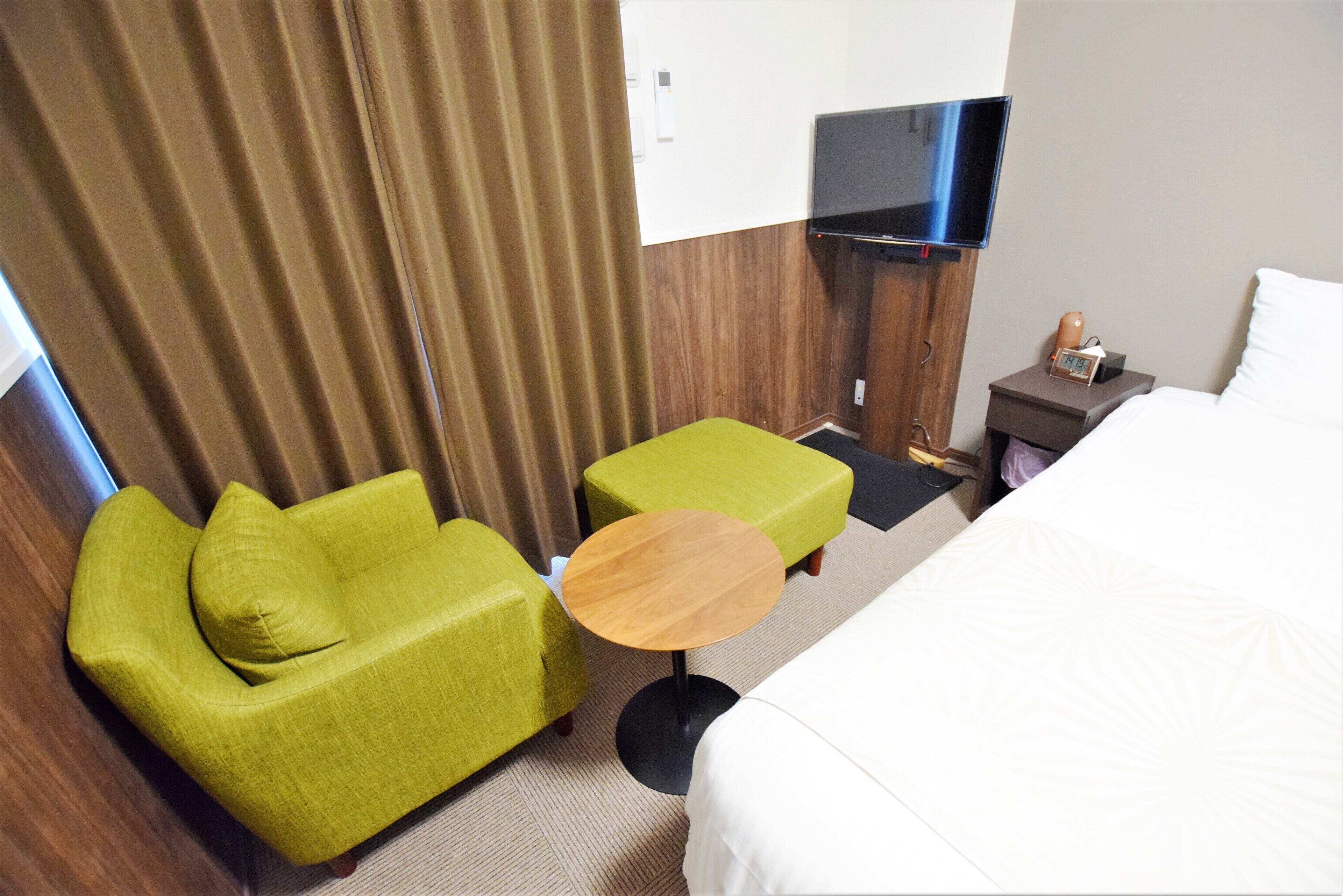A hotel room with a bed, a green armchair, a small wooden table, a TV mounted on the wall, and a nightstand with a lamp and a clock.