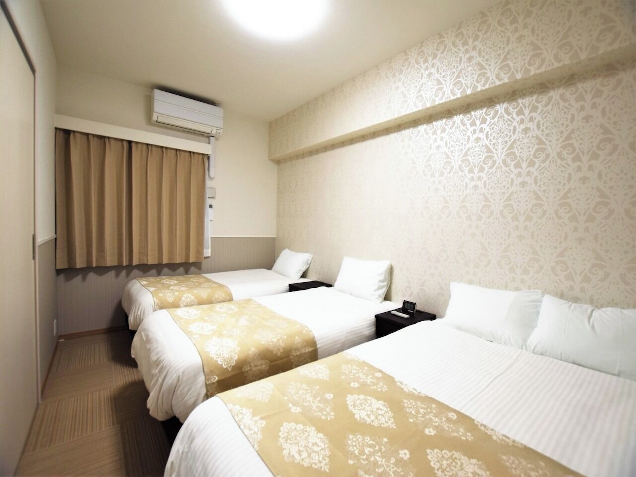 Ein Hotelzimmer mit drei Betten, einem fensterbelaubten Fenster, einer Klimaanlage und einer Wand mit Muster.