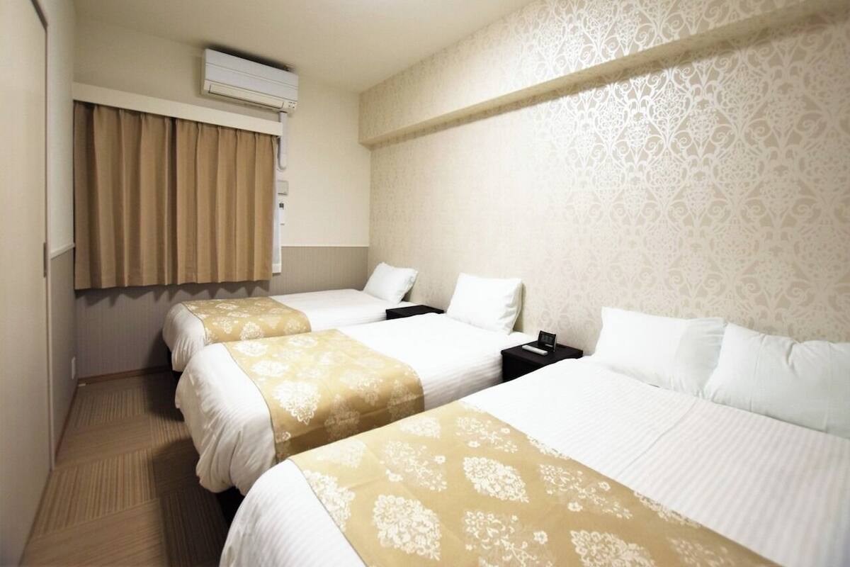 Ein Hotelzimmer mit drei Betten, einem fensterbelaubten Fenster, einer Klimaanlage und einer Wand mit Muster.