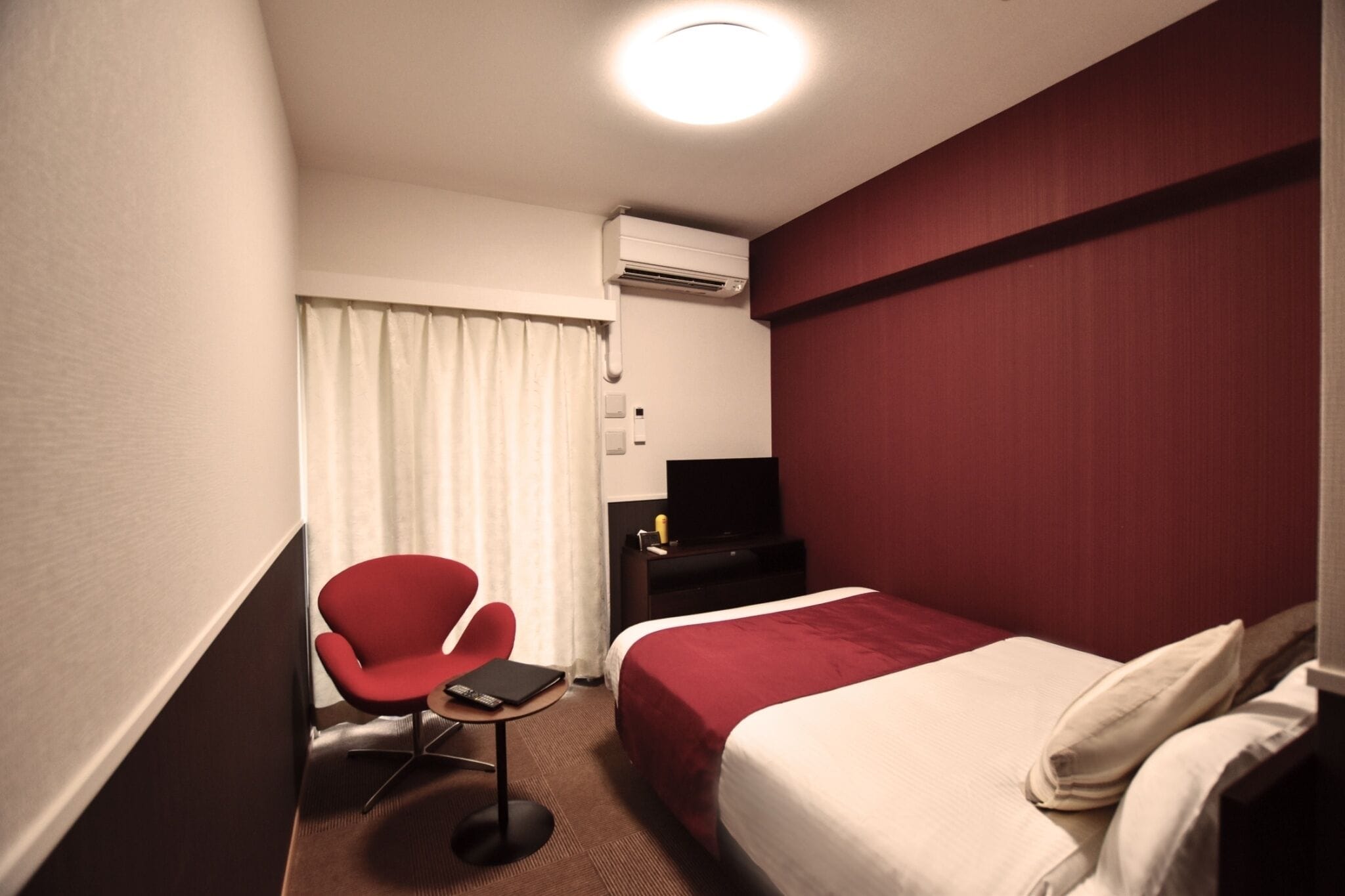 A hotel room with a bed, a red chair, a small table, an air conditioning unit, and a television on a black stand.
