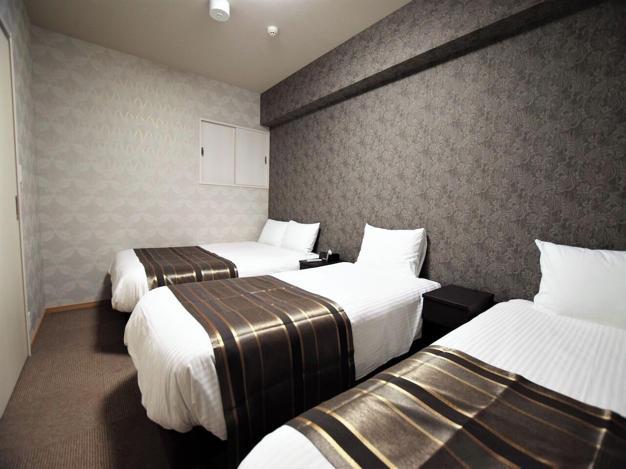 Ein Hotelzimmer mit drei Betten, einer grau gemusterten Wand und einer weißen Tür.