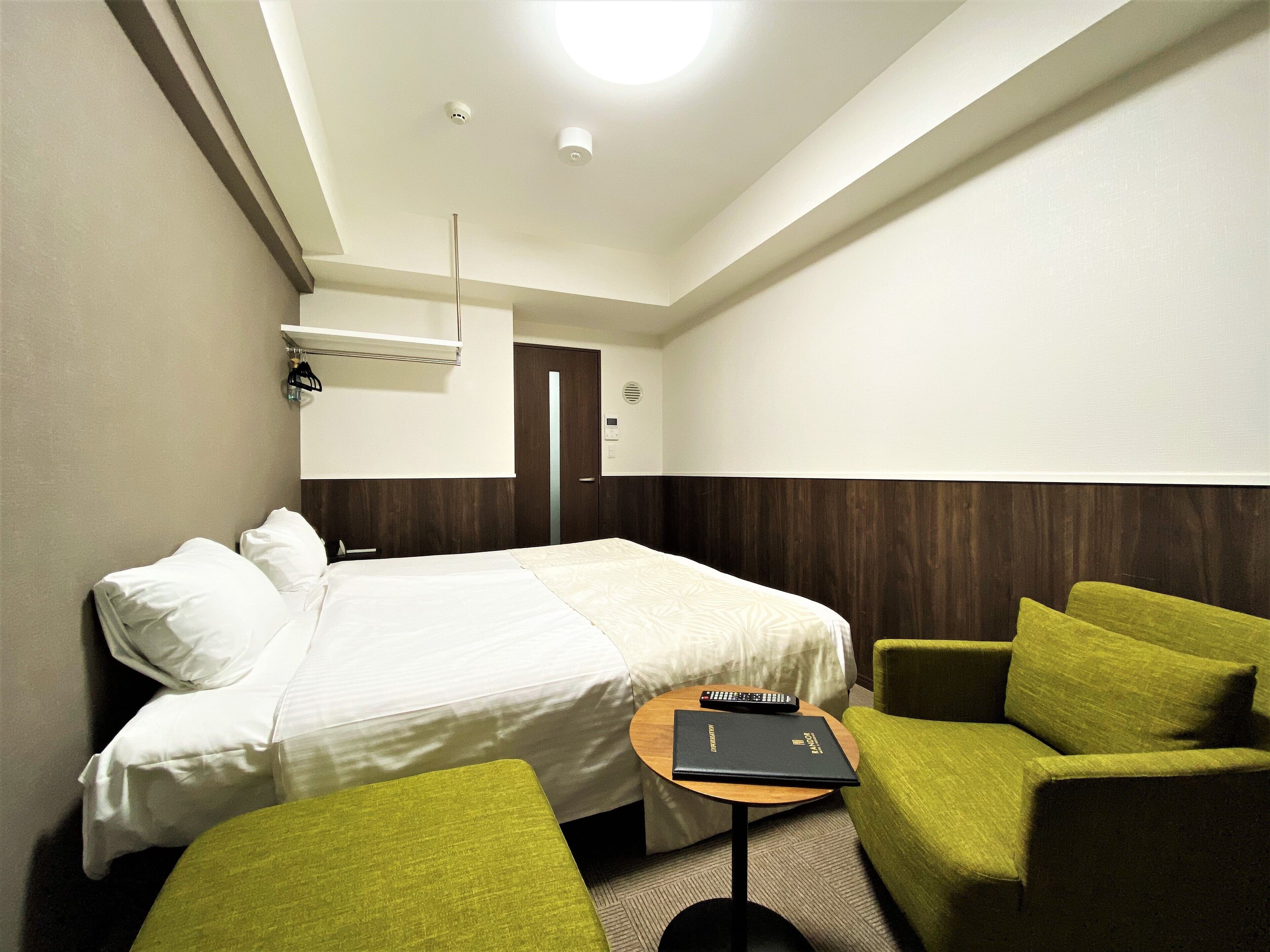 Una habitación de hotel con una cama, un sillón verde, una mesita con un libro y una puerta.