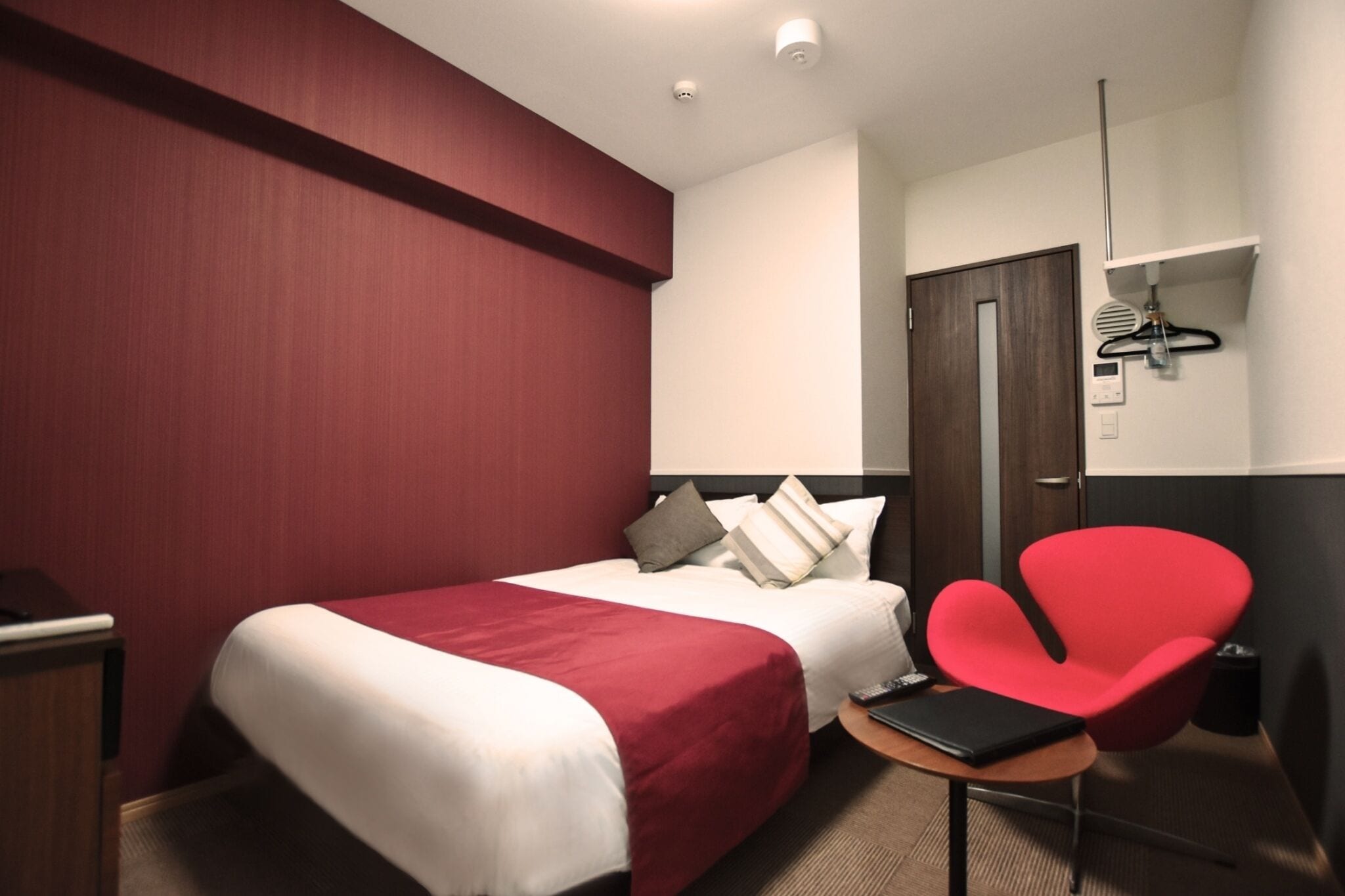 A hotel room with two beds, a red chair, a small table, and a wardrobe.