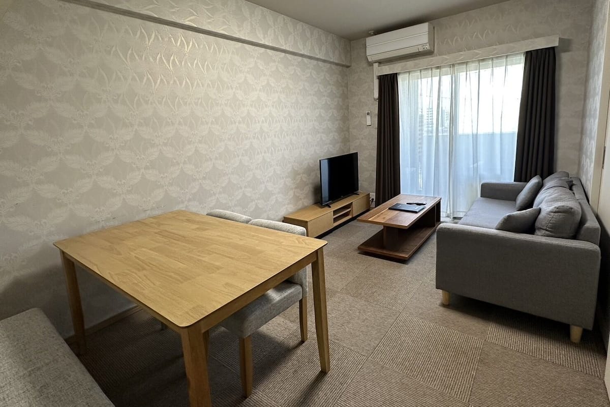 Ein modernes Wohnzimmer mit Esstisch aus Holz, einem grauen Sofa, einem Flachbildfernseher und einem großen Fenster mit Vorhängen.