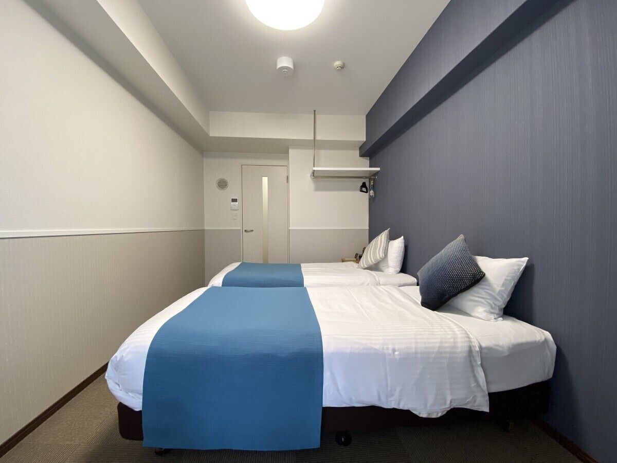 Ein Hotelzimmer mit zwei Betten, einem blauen Akzent an den Betten, einer weißen Tür und einer Leuchte an der Decke.