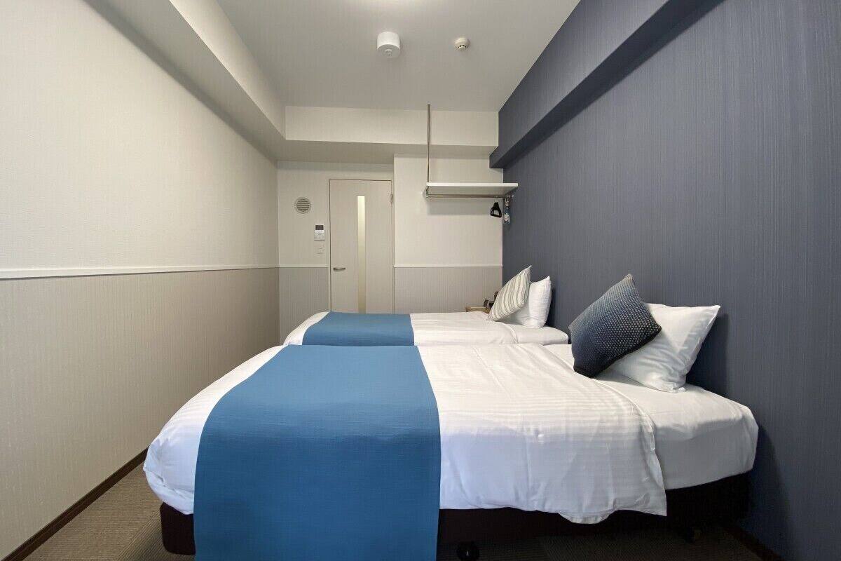 Ein Hotelzimmer mit zwei Betten, einem blauen Akzent an den Betten, einer weißen Tür und einer Leuchte an der Decke.