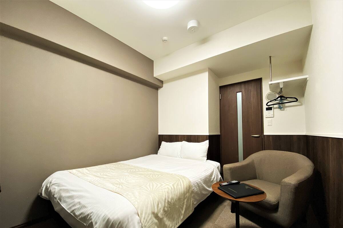Ein Hotelzimmer mit Bett, Stuhl, kleinem Tisch, Spiegel und Kleiderhaken.