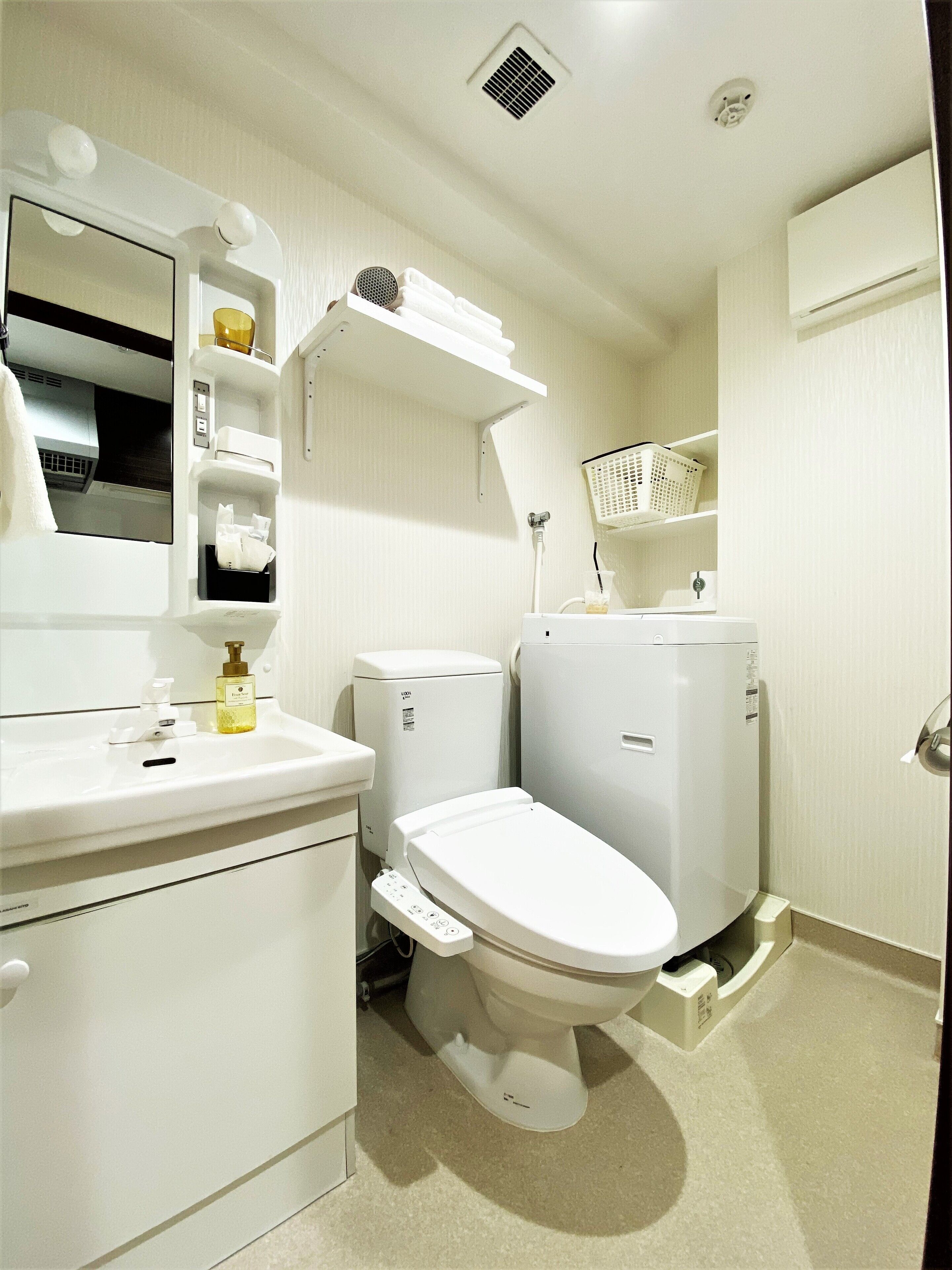 Une petite salle de bain propre, équipée de toilettes, d’un lavabo et d’un miroir. On y trouve des étagères au-dessus du lavabo et un panier fixé au mur.