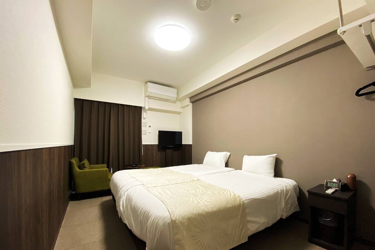 Ein Hotelzimmer mit einem großen Bett, einem an der Wand befestigten Fernseher, einem kleinen Tisch mit einer Uhr und einem grünen Sessel.