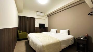 A hotel room with a large bed, a television on the wall, a small table with a clock, and a green chair.