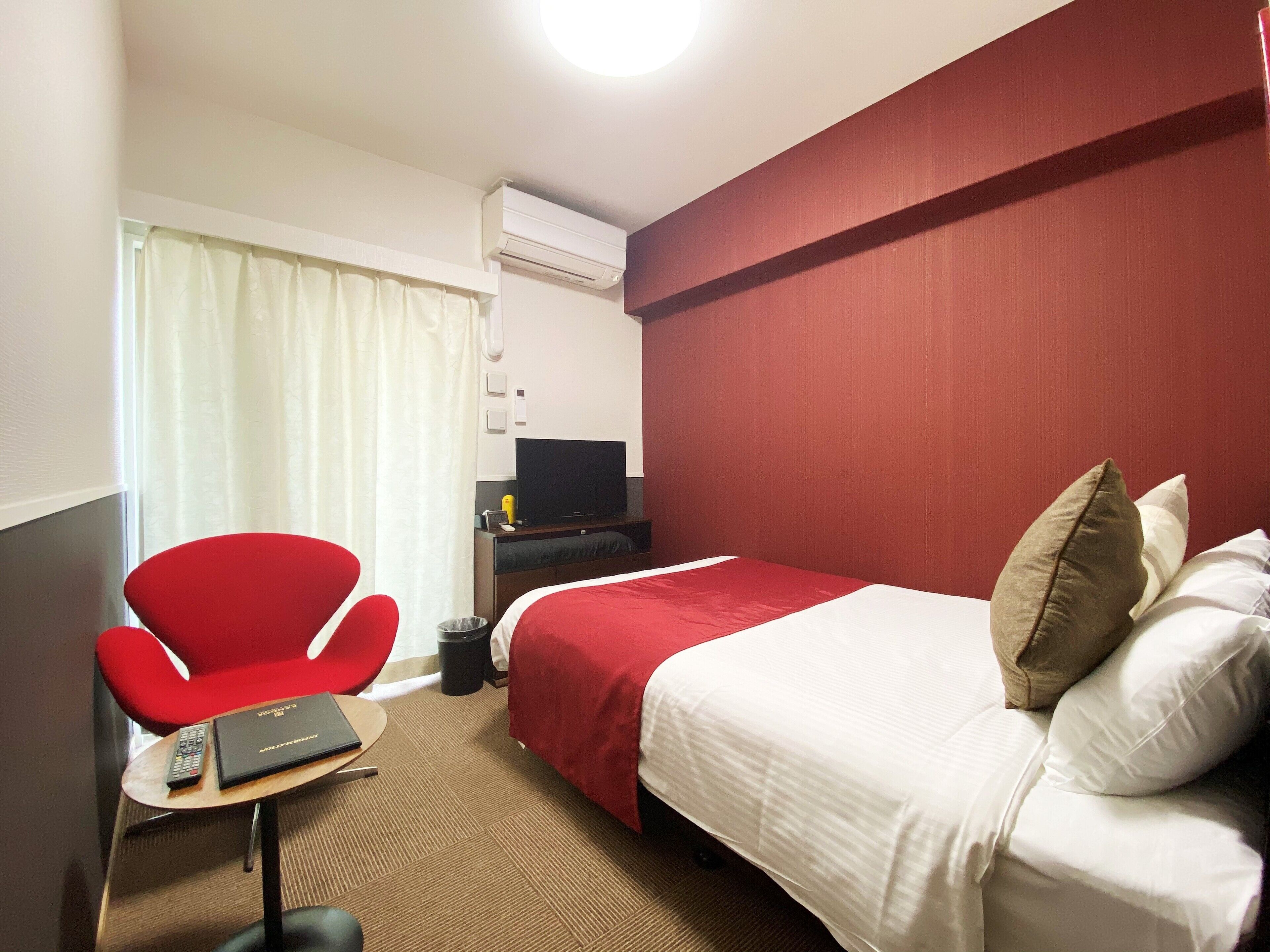 Ein Hotelzimmer mit einem Bett, einem roten Sessel, einem kleinen Tisch mit Fernbedienung, einem Fernseher und einer Klimaanlage.