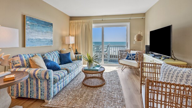 A spacious living room with a sofa, a coffee table, and a television. A balcony with a view of the ocean is visible. - Sea Cloisters II Ocean Front Beach Retreat (North Myrtle Beach)