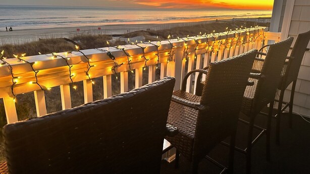 A balcony with wicker chairs, a railing adorned with string lights, and a view of the ocean at sunset. - Sea Cloisters I Ocean Front Beach Retreat (North Myrtle Beach)