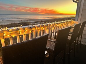 Un balcón con sillas de mimbre, barandilla decorada con luces de adorno y vista al océano al atardecer.