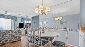A dining area with a round table, white chairs, and a chandelier. There is a sofa with patterned upholstery and a large window with curtains. - Sea Cloisters I Ocean Front Beach Retreat (North Myrtle Beach)
