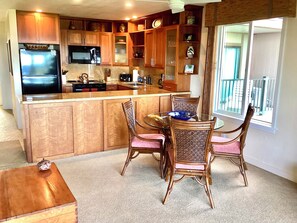 A kitchen with wooden cabinets, a refrigerator, a microwave, and a dining area with wicker chairs and a glass table. - It's all About the View! Oceanfront, A/C, Elevator (Kapaa)