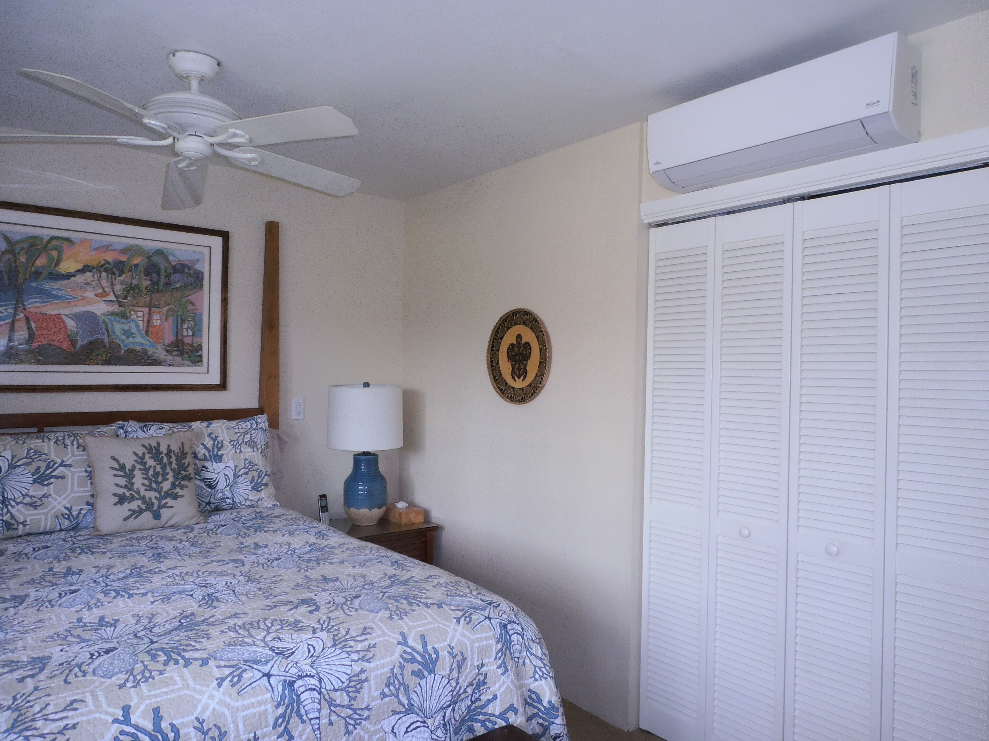 A bedroom with a bed, a ceiling fan, a wall-mounted air conditioner, a bedside lamp, a framed picture, and a closet with louvered doors.