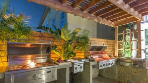 An outdoor kitchen with a grill, a sink, and a stove, surrounded by plants and a pergola. - The Self-Sustainable Beach House (South Padre Island)