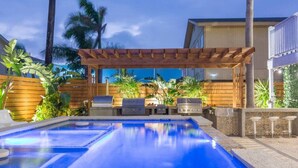 A swimming pool with a wooden pergola overhead, surrounded by tropical plants and a barbecue area. - The Self-Sustainable Beach House (South Padre Island)