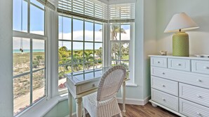 A room with a desk and chair facing large windows with a view of the beach and palm trees.