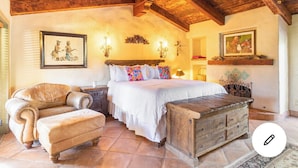 Un dormitorio con techo de madera, una cama con ropa de cama blanca, un sillón de cuero y un aparador de madera.