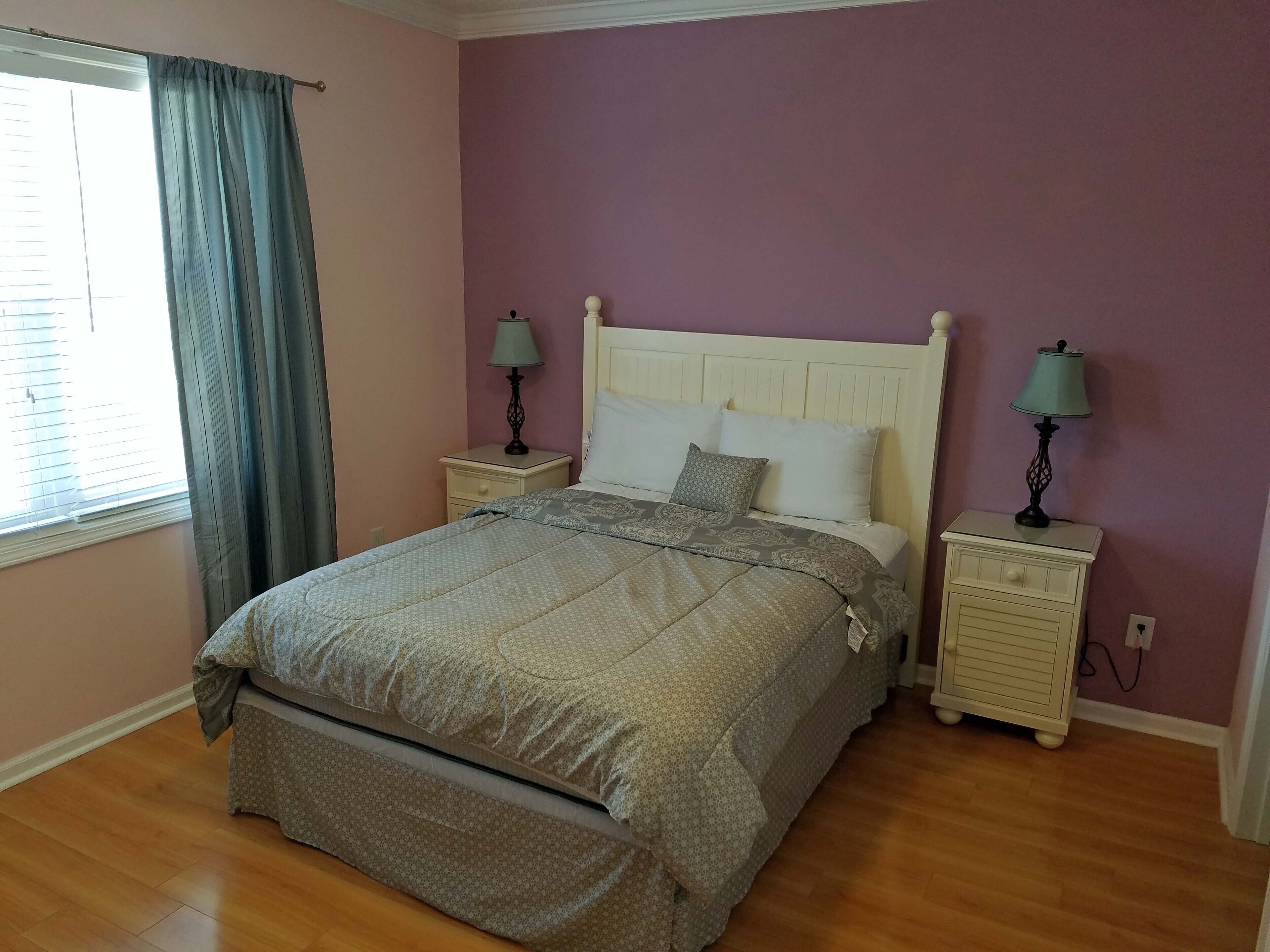 A bedroom with a bed, two bedside tables with lamps, a window with curtains, and a wooden floor.